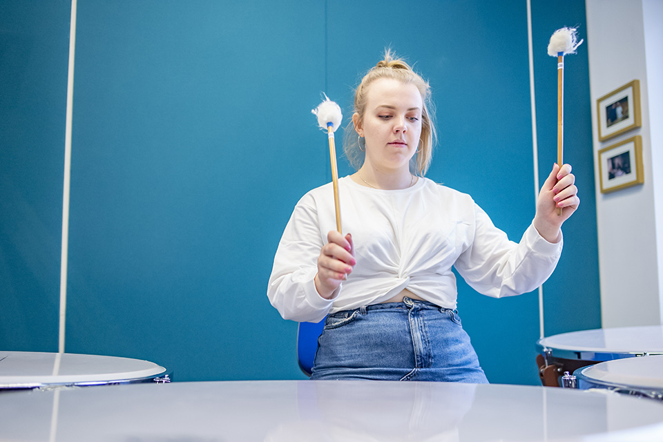 A female student performing on a timpani drum with some mallets, with a blue background.